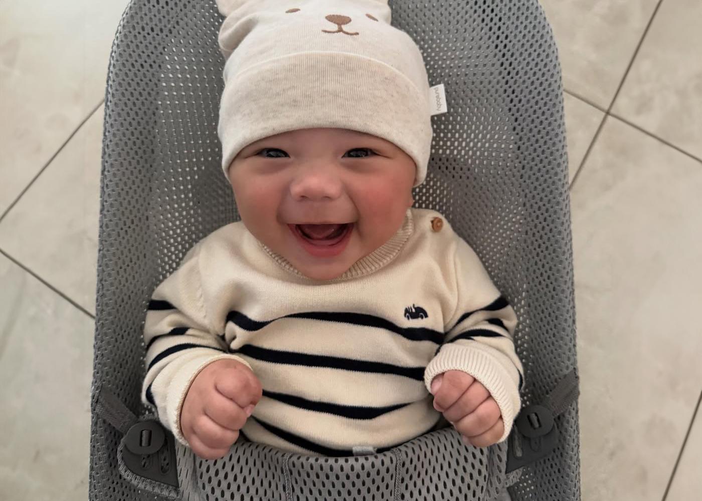 Smiling baby wearing a striped sweater and bear-themed hat, sitting in a gray mesh baby seat on a tiled floor.