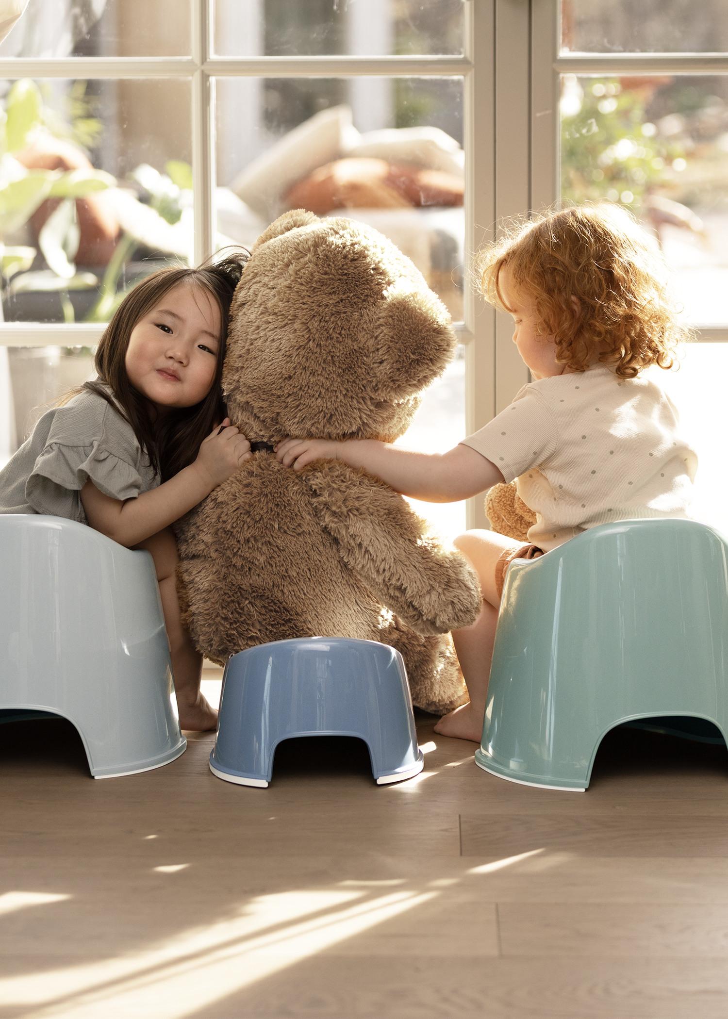 Two children sit on stools, hugging a large teddy bear in a sunlit room with a glass door.