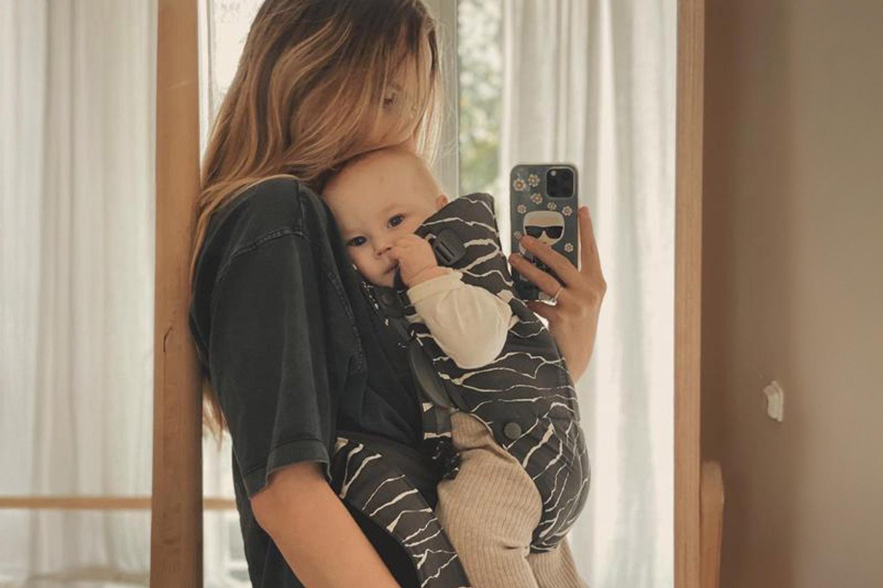 Person taking a mirror selfie while holding a baby in a patterned carrier against sheer curtains.