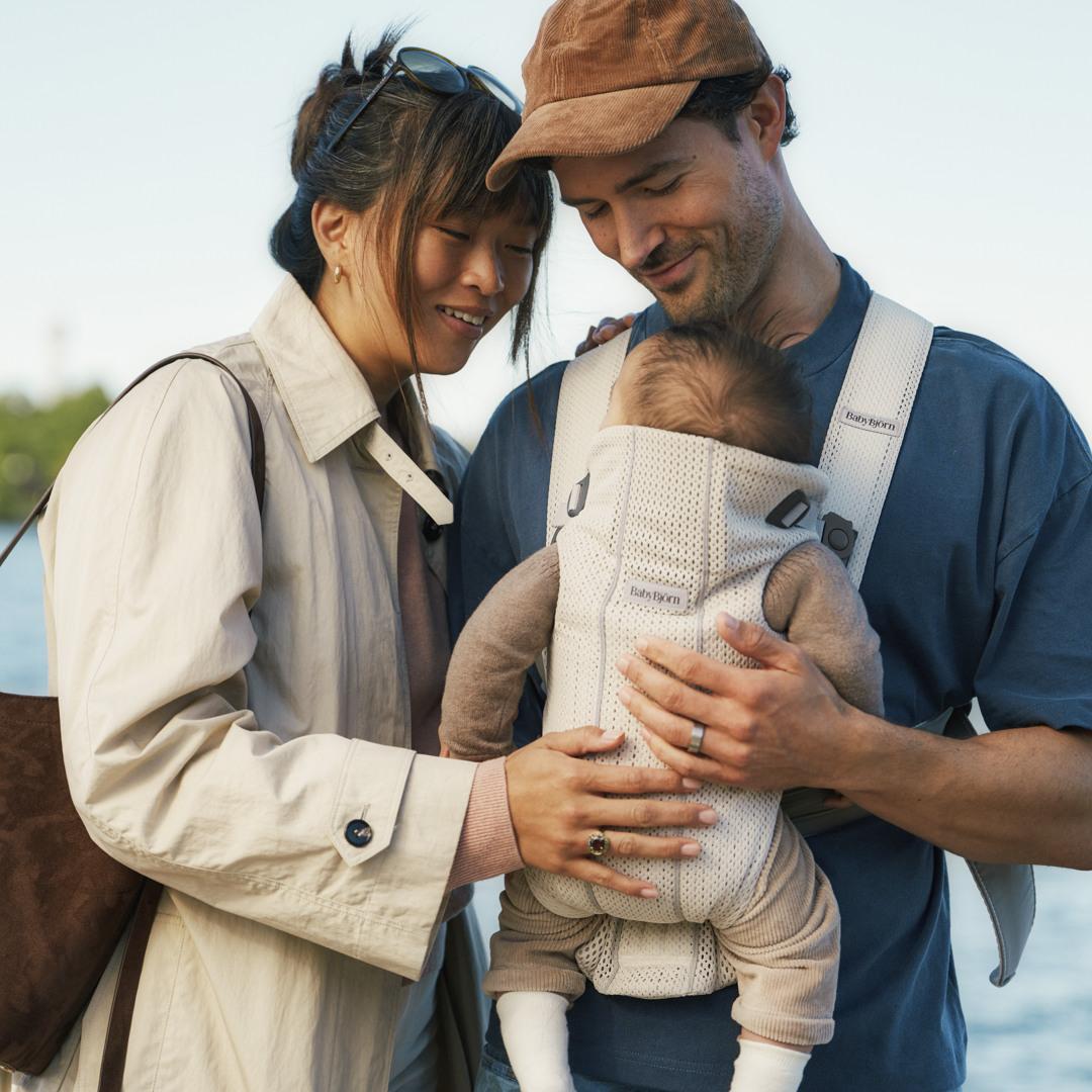 A happy couple stands outside, smiling at their baby in a front carrier. The woman wears a beige coat, and the man wears a cap.