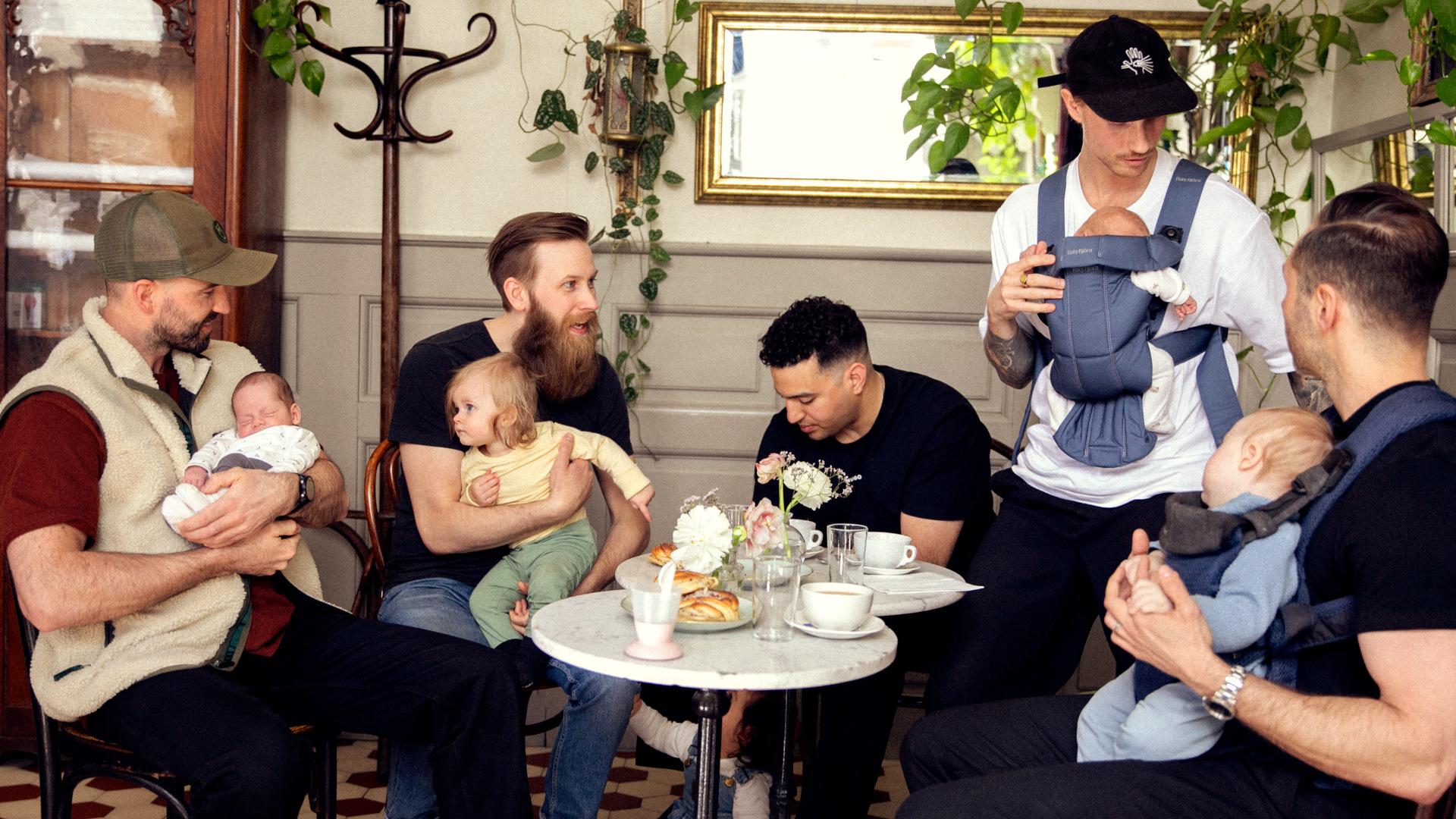 Group of five men with babies, sitting and standing around a café table, surrounded by plants and casual decor.