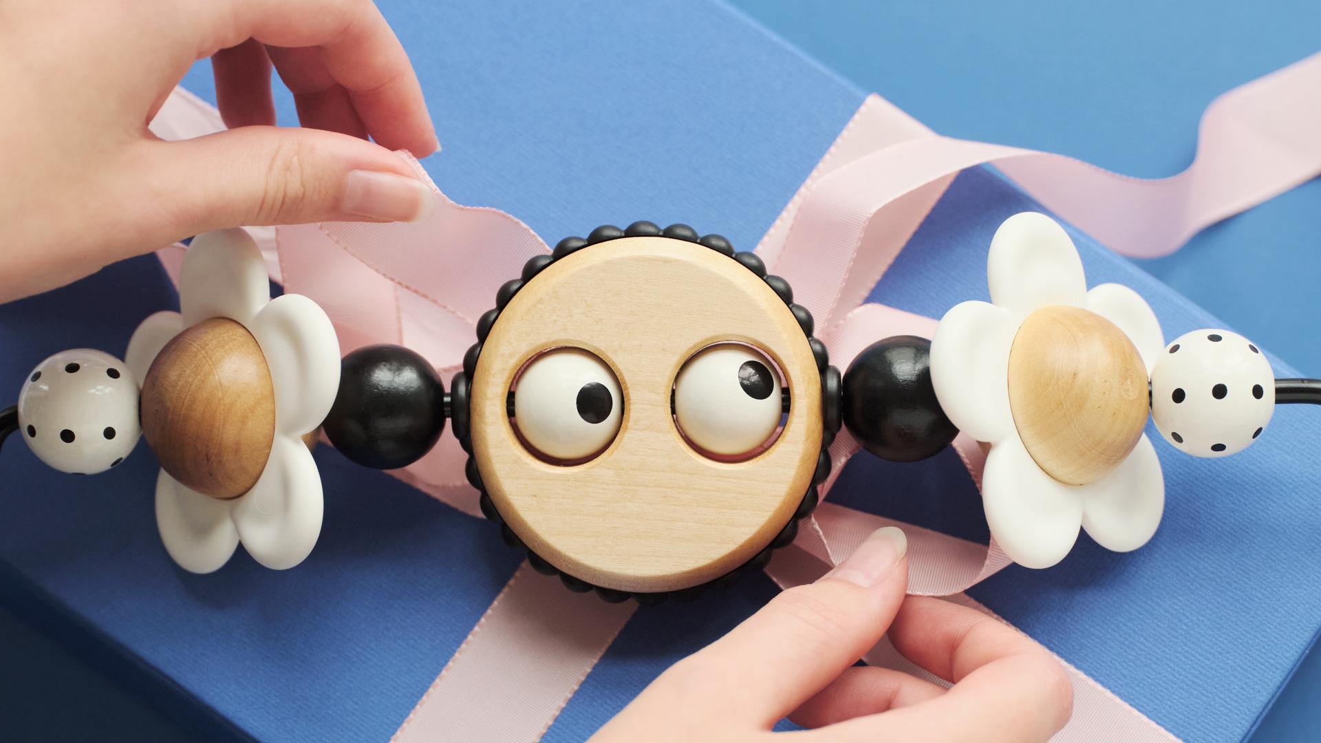 Hands holding a wooden toy with googly eyes, flanked by flower shapes and polka-dot spheres, on a blue gift box with a pink ribbon.