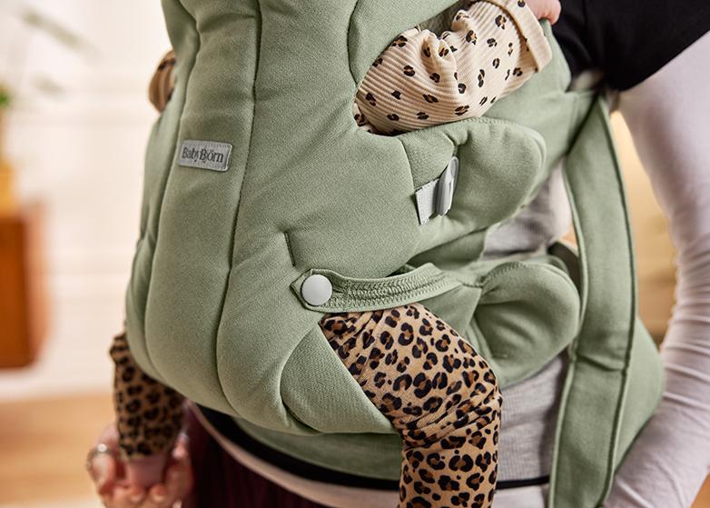 A baby wearing leopard print clothing is snugly secured in a green baby carrier.